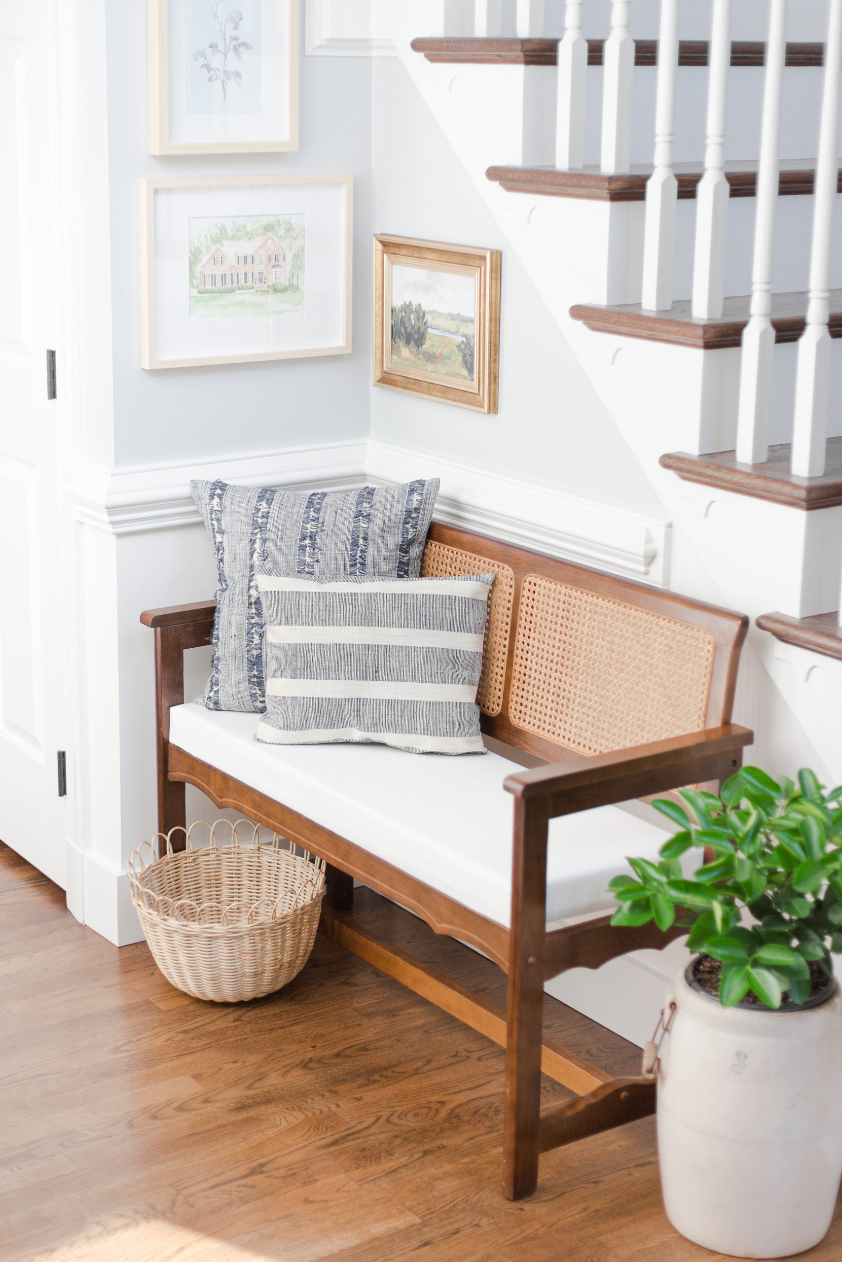 Entryway with cane bench, pillows, next to a staircase with green plant in pot on floor next to bench