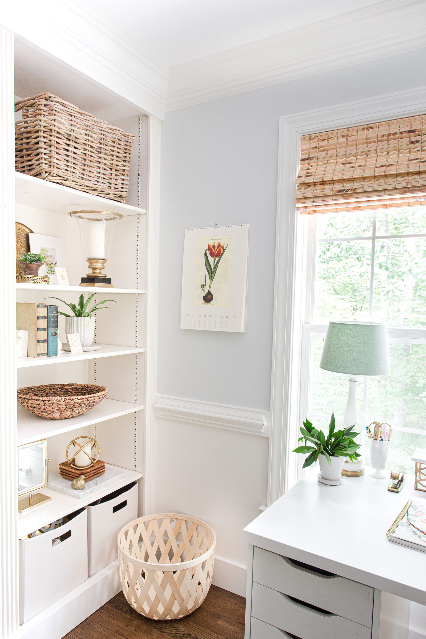 Bookcase in home office next to a desk overlooking a window with baskets on top shelf, lamp and green plant on desk