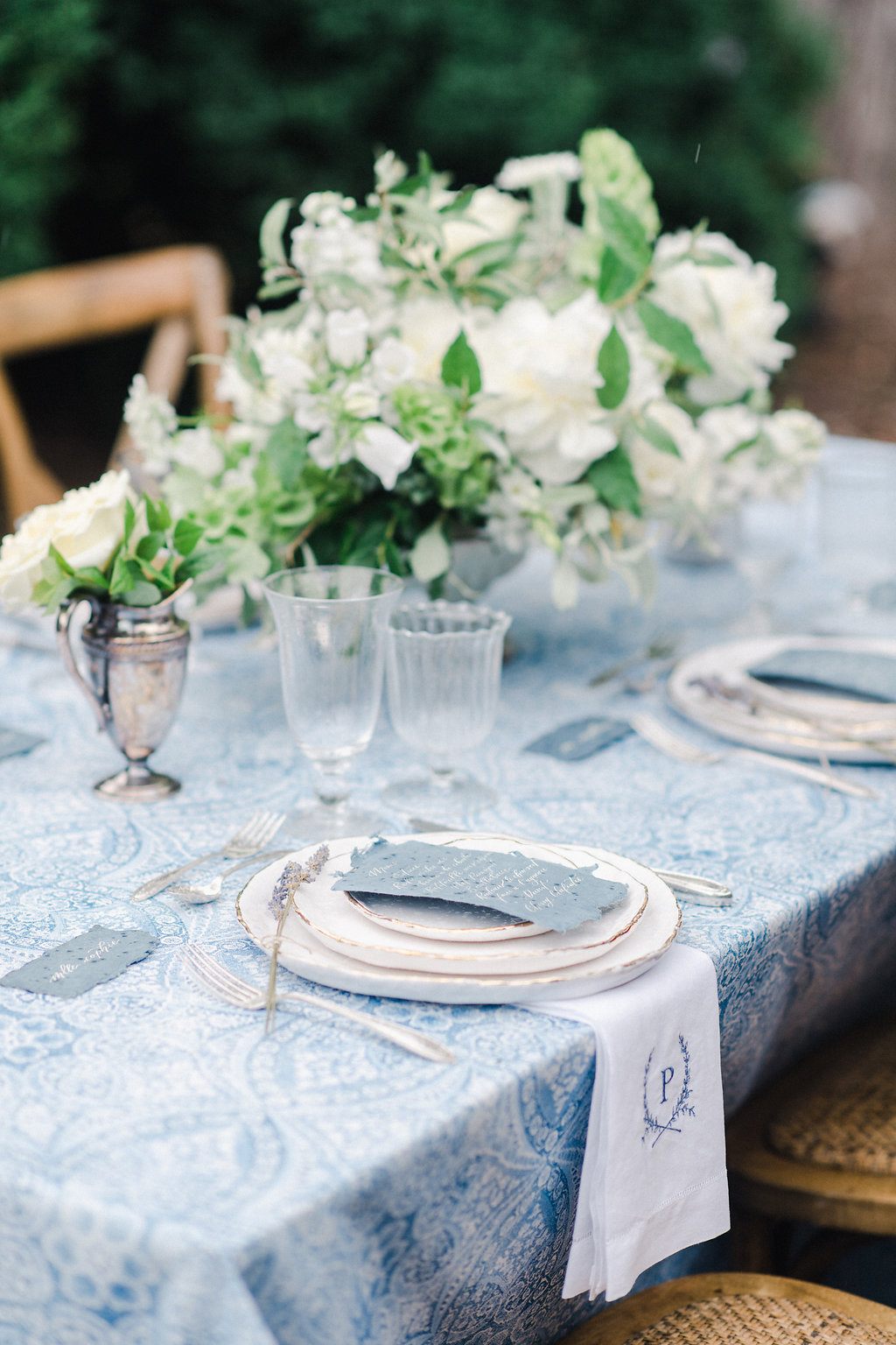Blue and white table cloth with dinner plates, glasses and white floral centerpiece outside. 