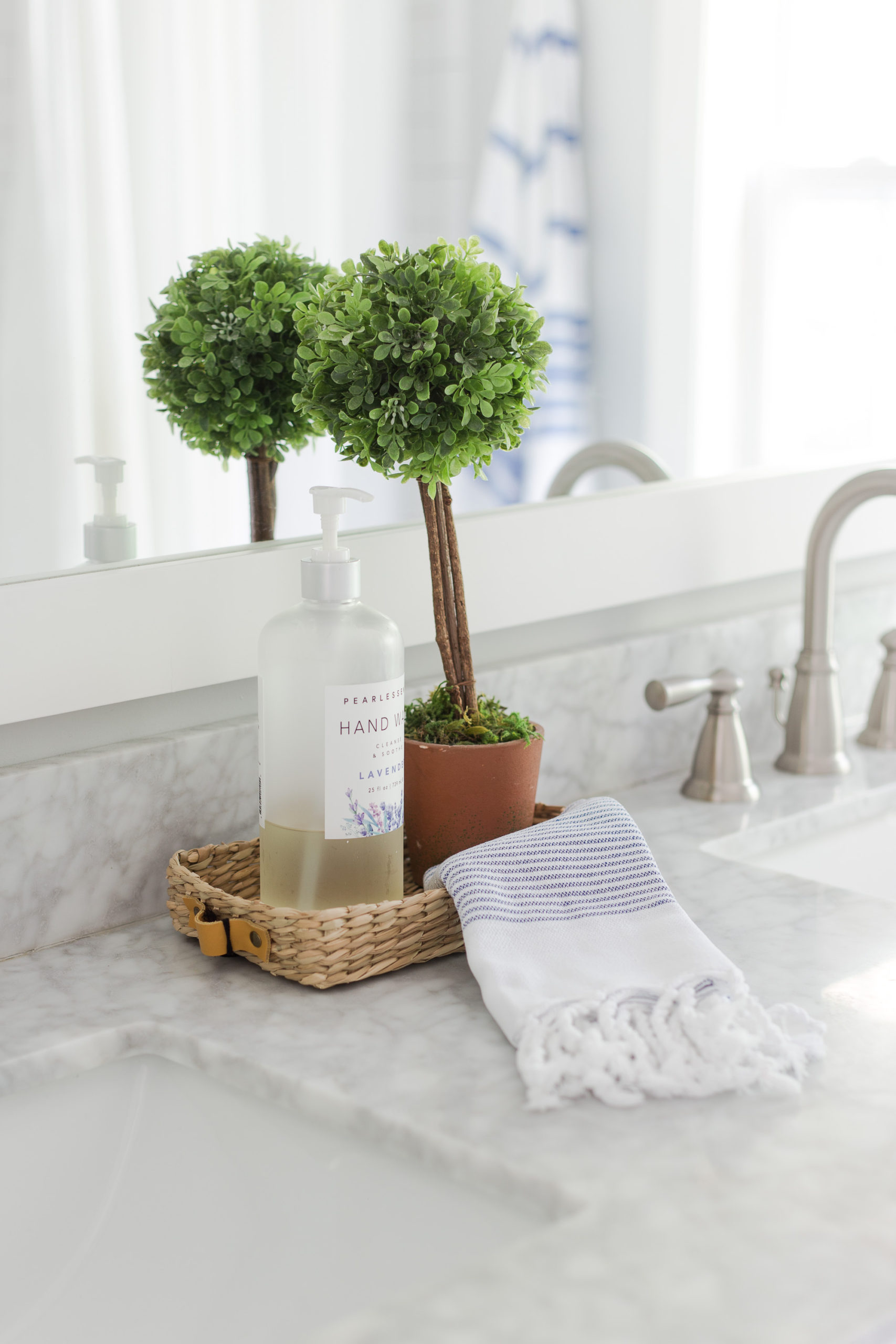 Jar of hand soap, and green topiary sitting in a small rattan tray with towel sitting on a bathroom countertop