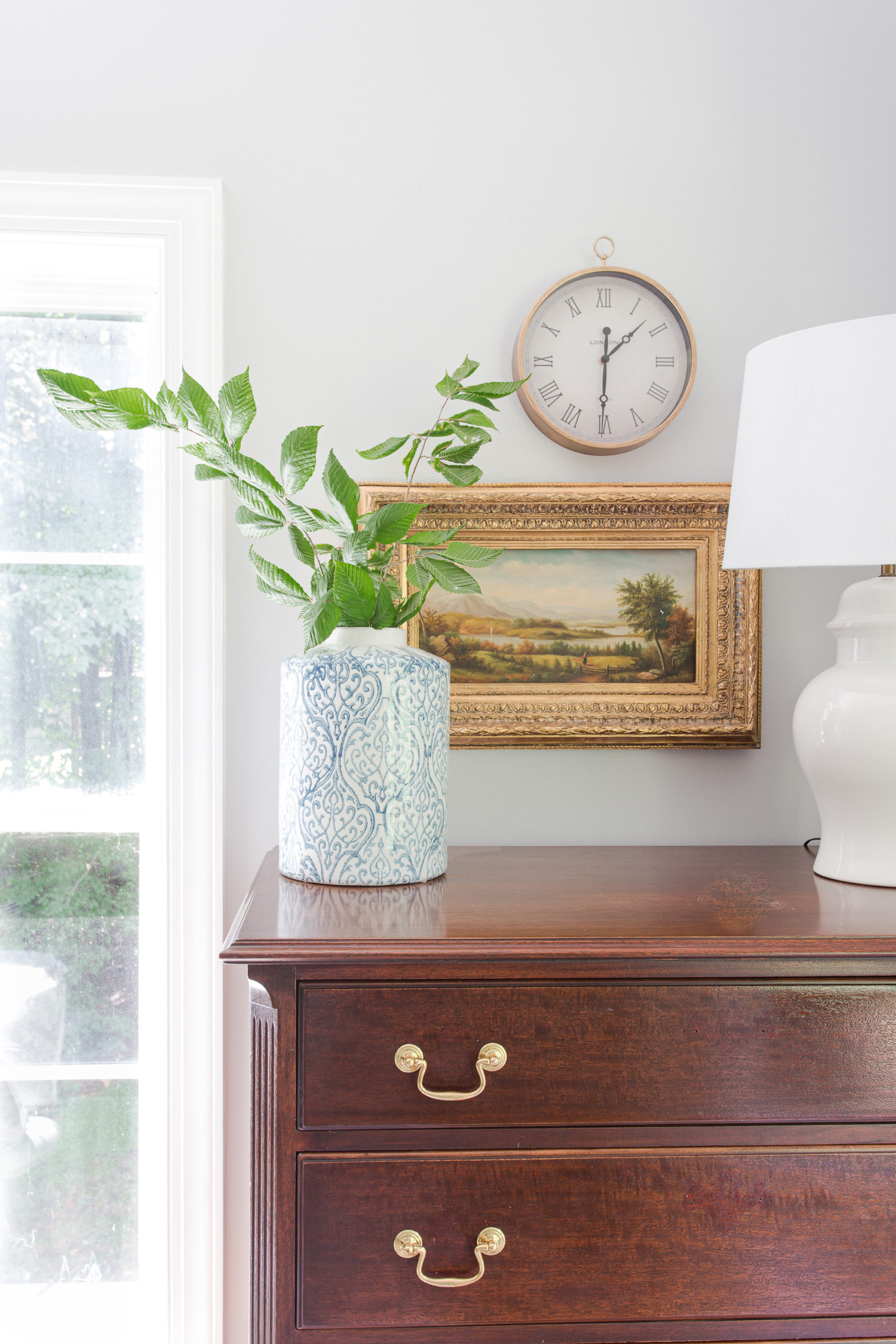 Dresser with blue and white vase and greenery sitting next to white lamp and clock hanging on the wall. 