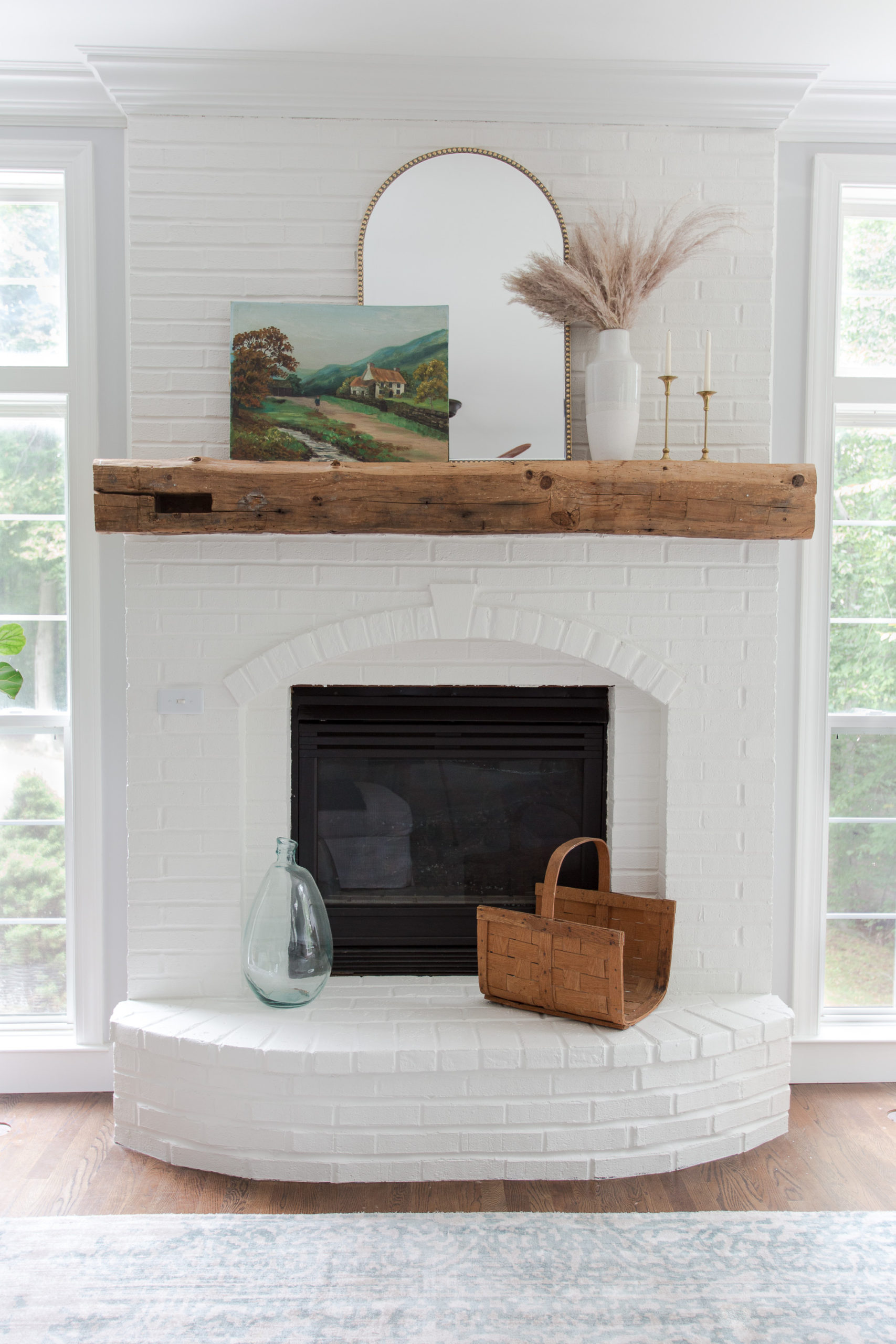 white brick fireplace with arched gold mirror and vintage painting propped on a wooden mantel 