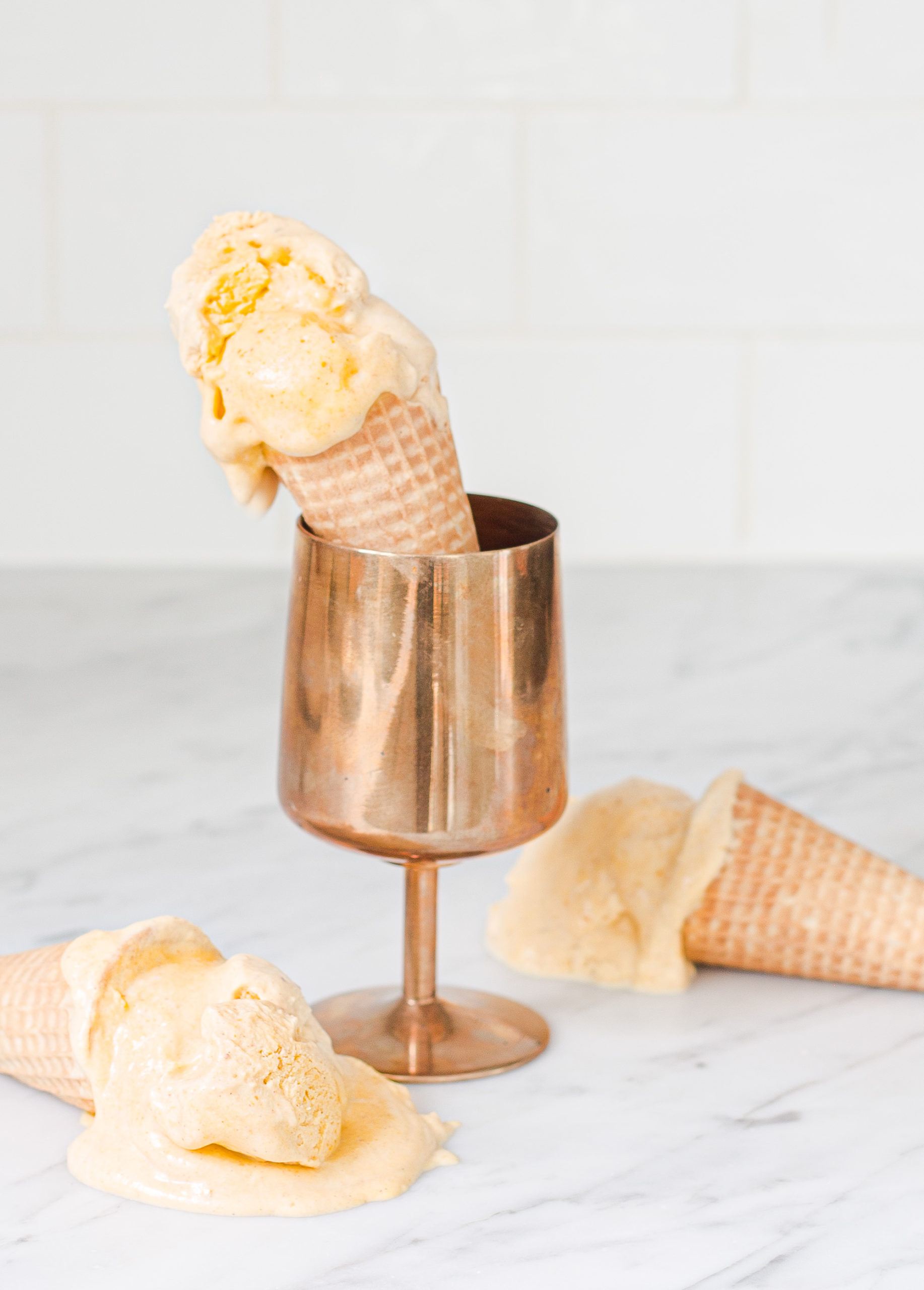 ice cream cone with ice cream propped up in copper goblet with two ice cream cones and melted ice cream laying on counter