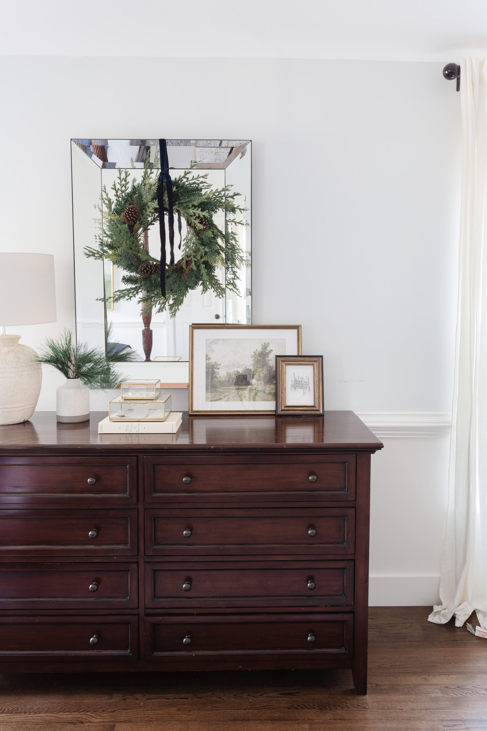 mirror hanging above dark wood dresser with greenery and artwork propped at base of mirror. 