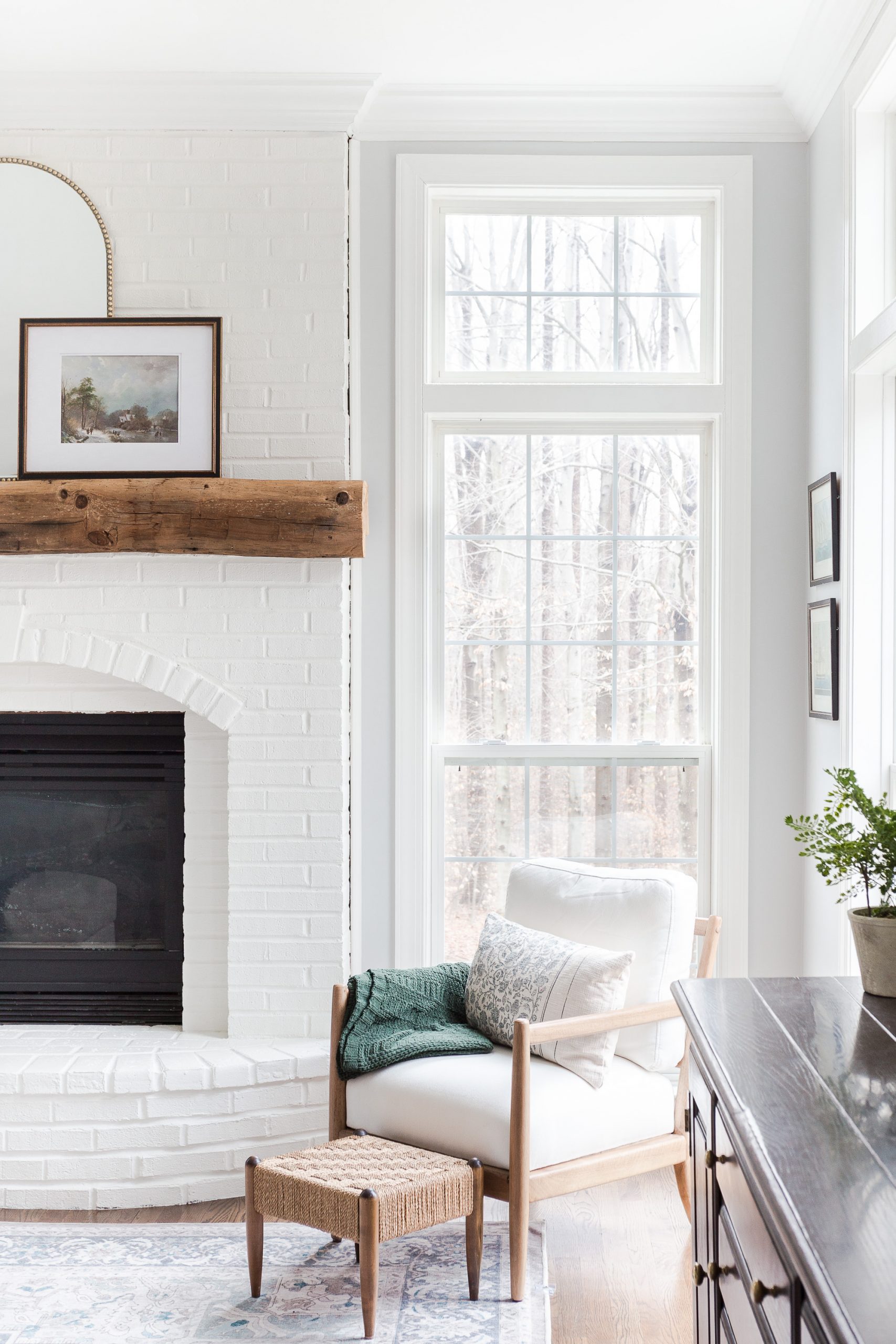 wooden chair with white cushions sitting between two windows next to a white brick painted fireplace