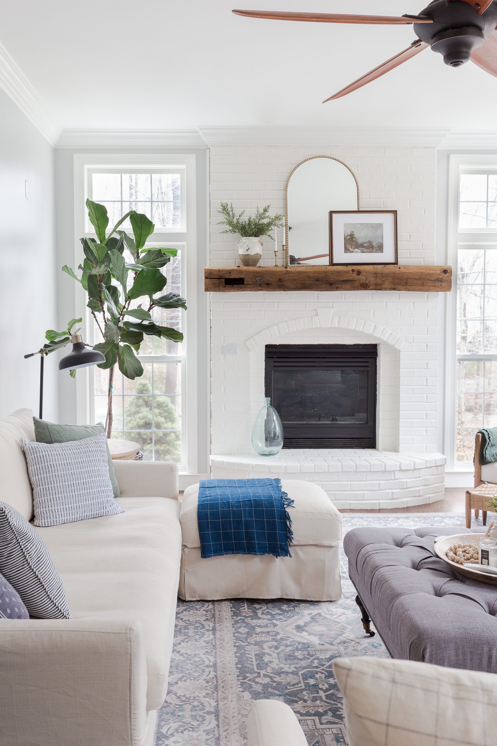 family room with painted white brick fireplace with fiddle leaf tree next to window in corner and sofa and gray tufted ottoman