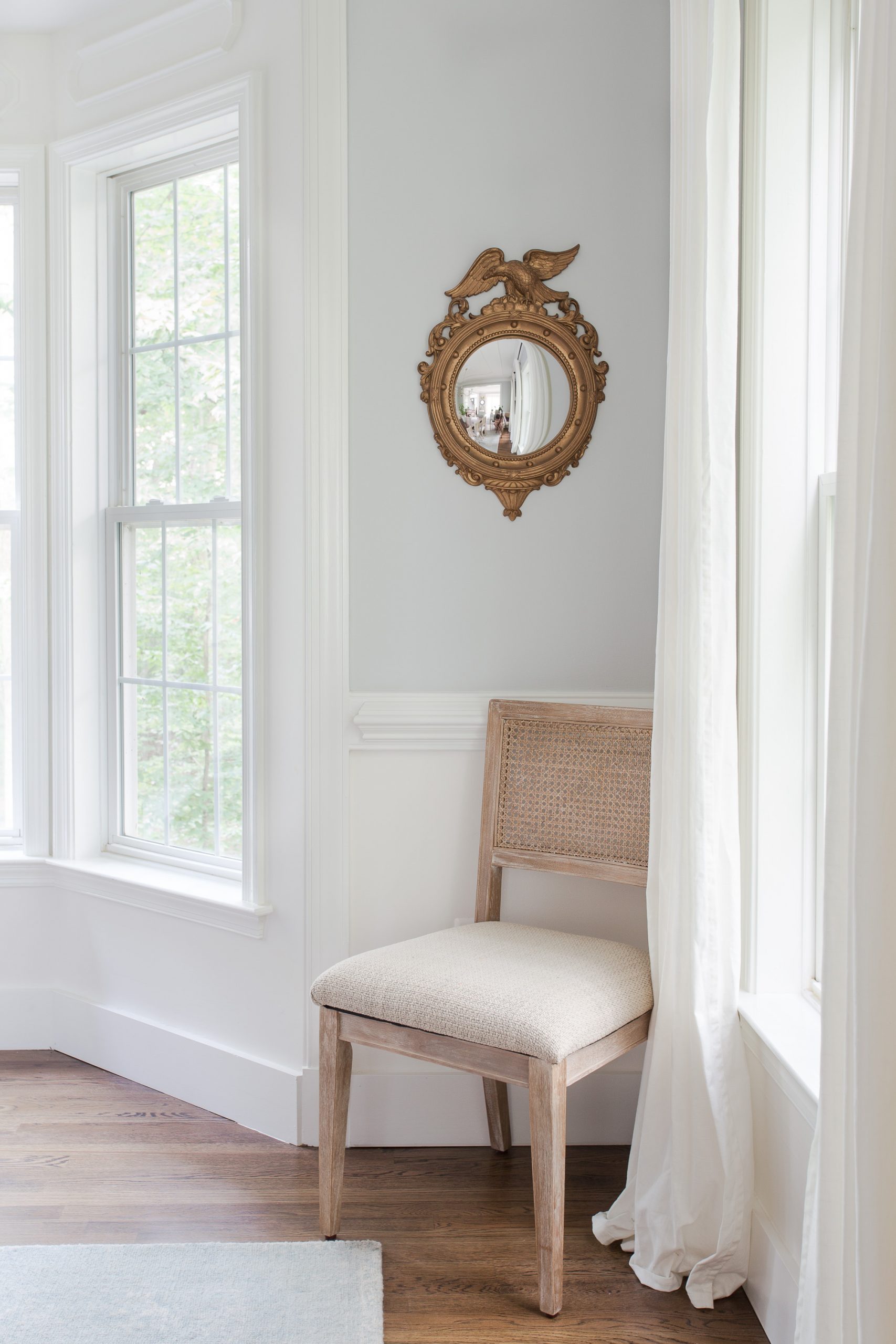 cane back chair in the corner of the dining room next to window with round gold mirror above chair