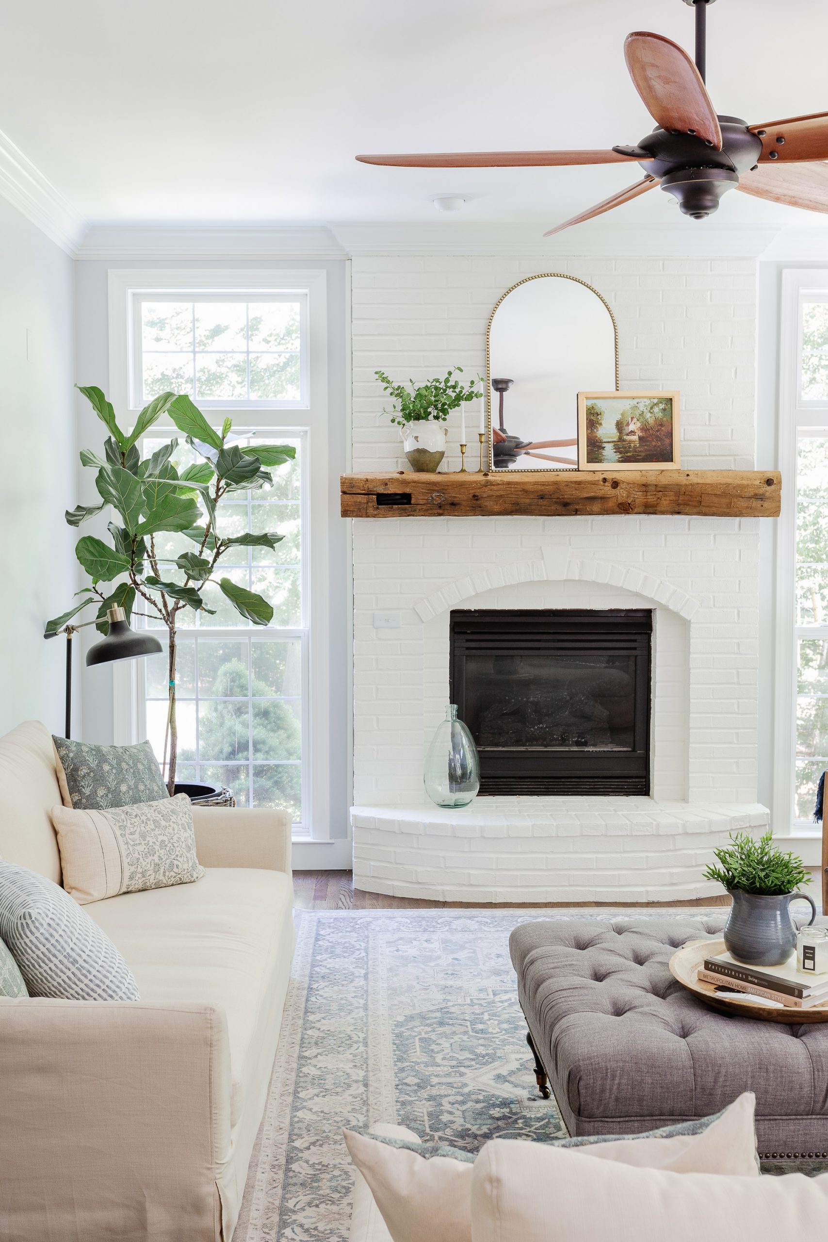 painted white brick fireplace in family room with ceiling fan, sofa, fiddle leaf tree next to window in corner. 