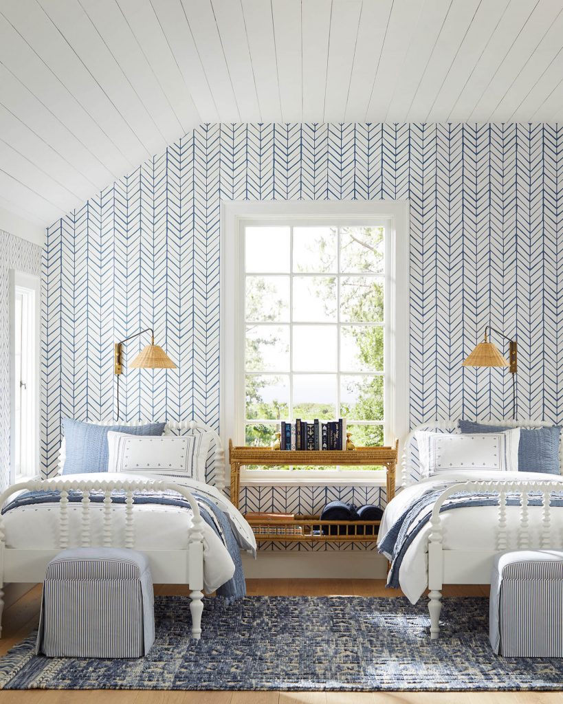 Blue and white wall paper on wall with two white spindles beds and window in between the beds. 