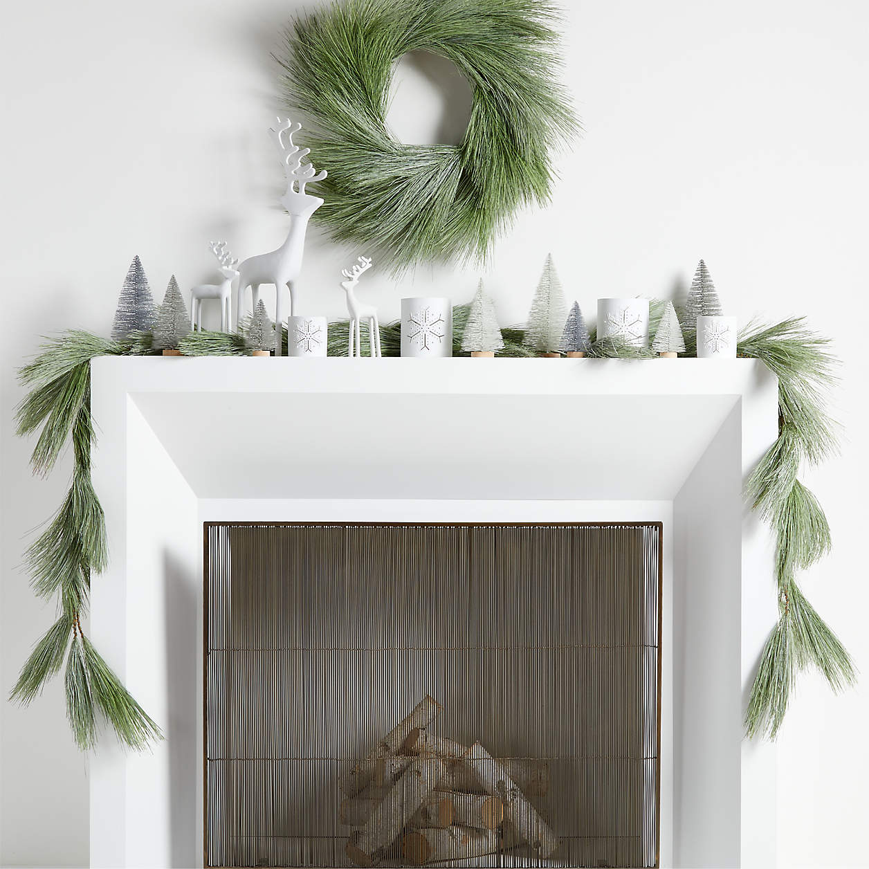 white fireplace with green pine garland, wreath hanging above and little christmas trees on the mantel
