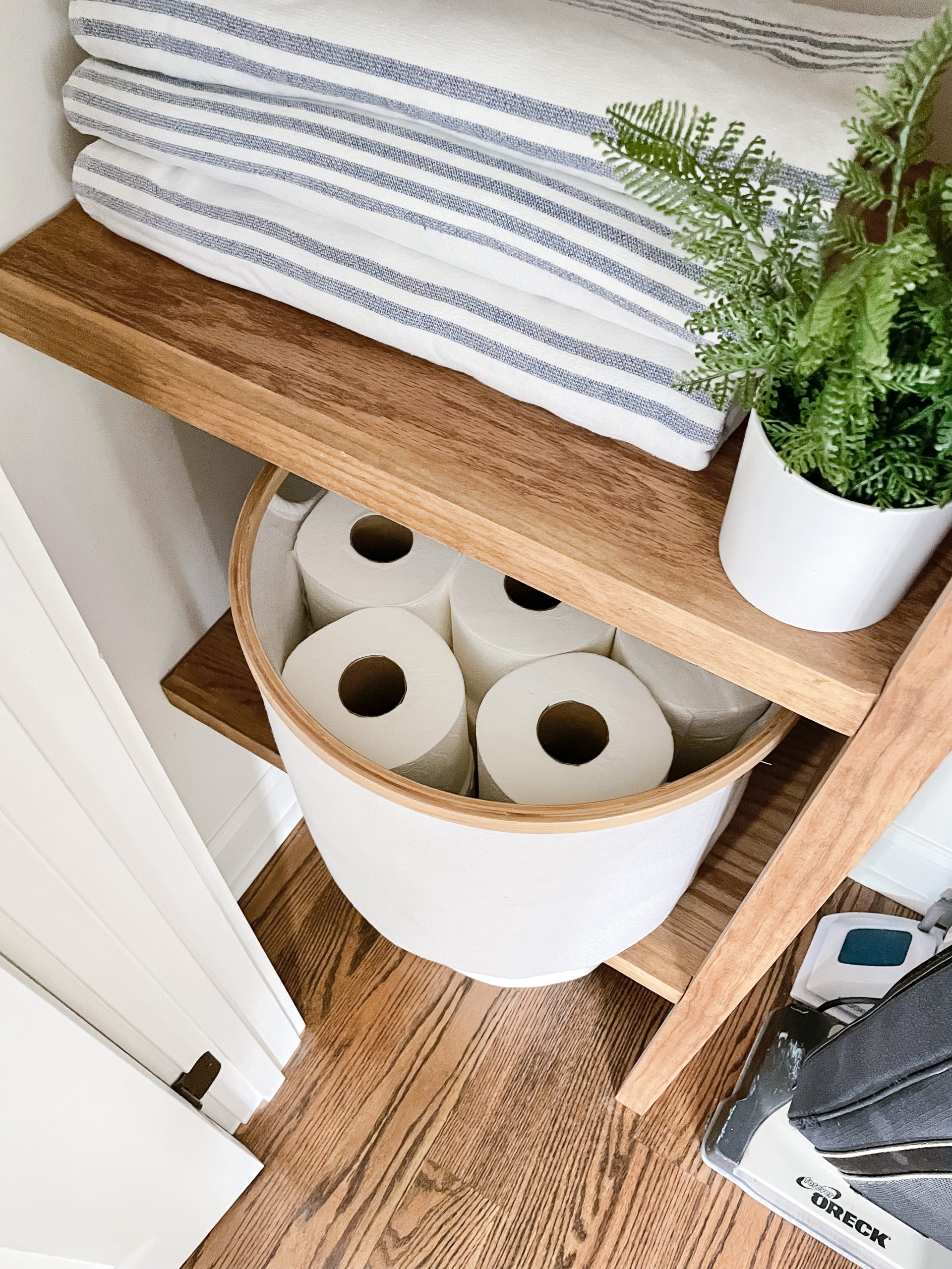 bin of toilet paper on wood shelf and blue and white stripe towel and plant on another shelf.