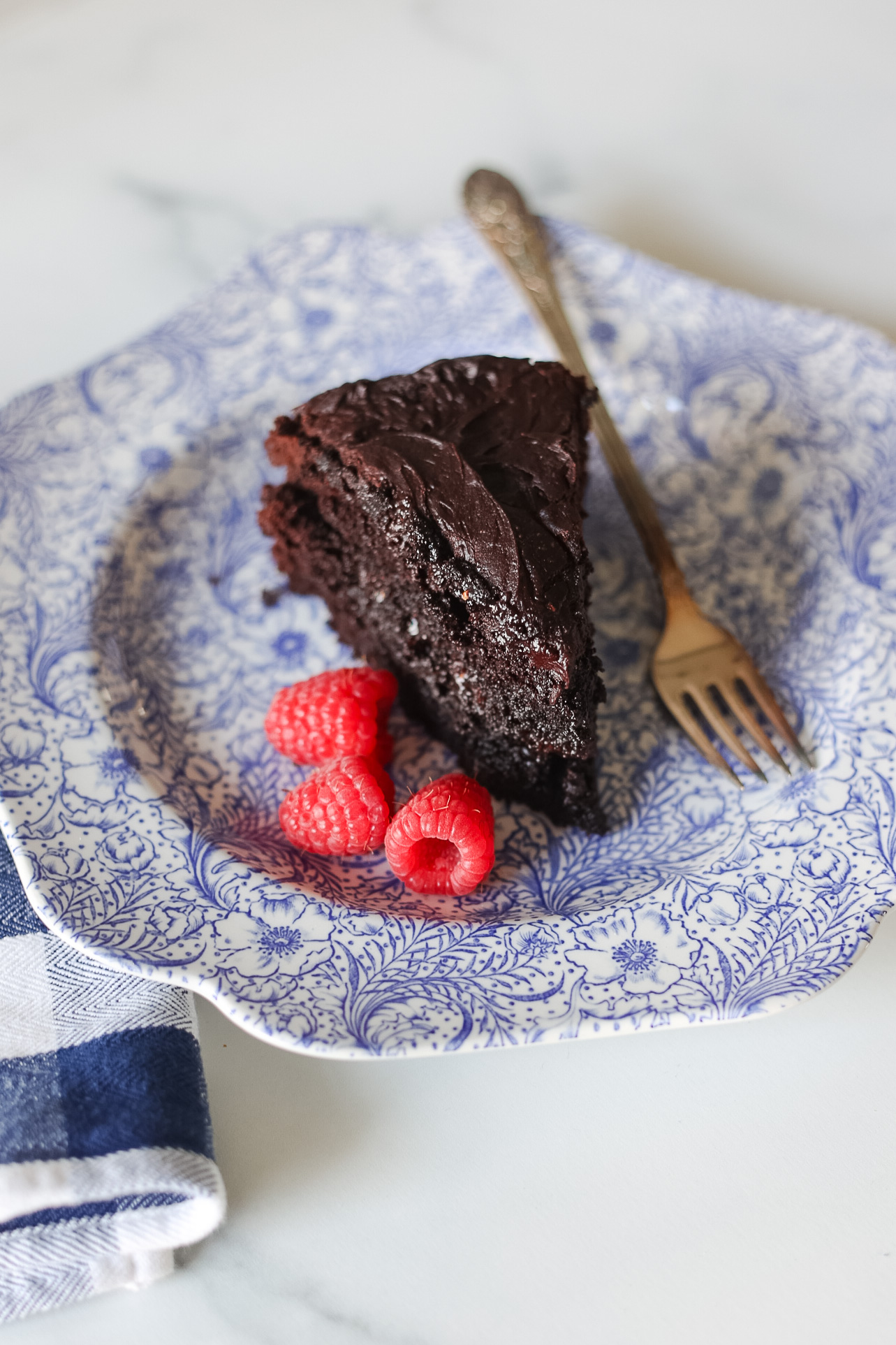 dark chocolate cake slice on blue and white floral plate with raspberries on plate