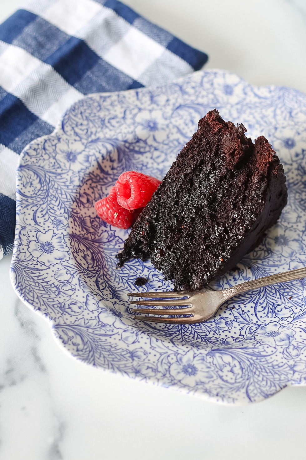 dark chocolate cake slice on blue and white floral plate with raspberries on plate