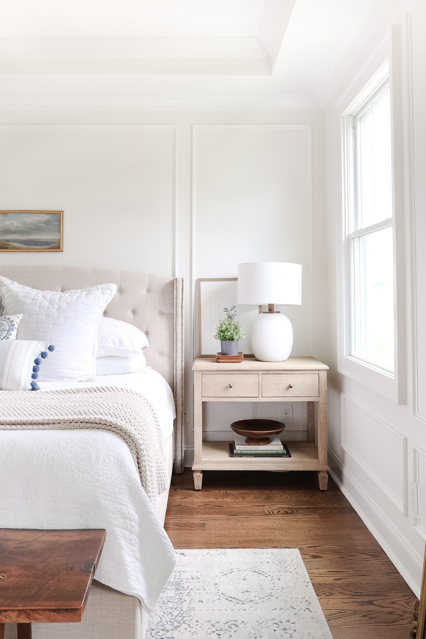White bedroom with pvc trim bed with nightstand next to it and lamp and plant on top. 