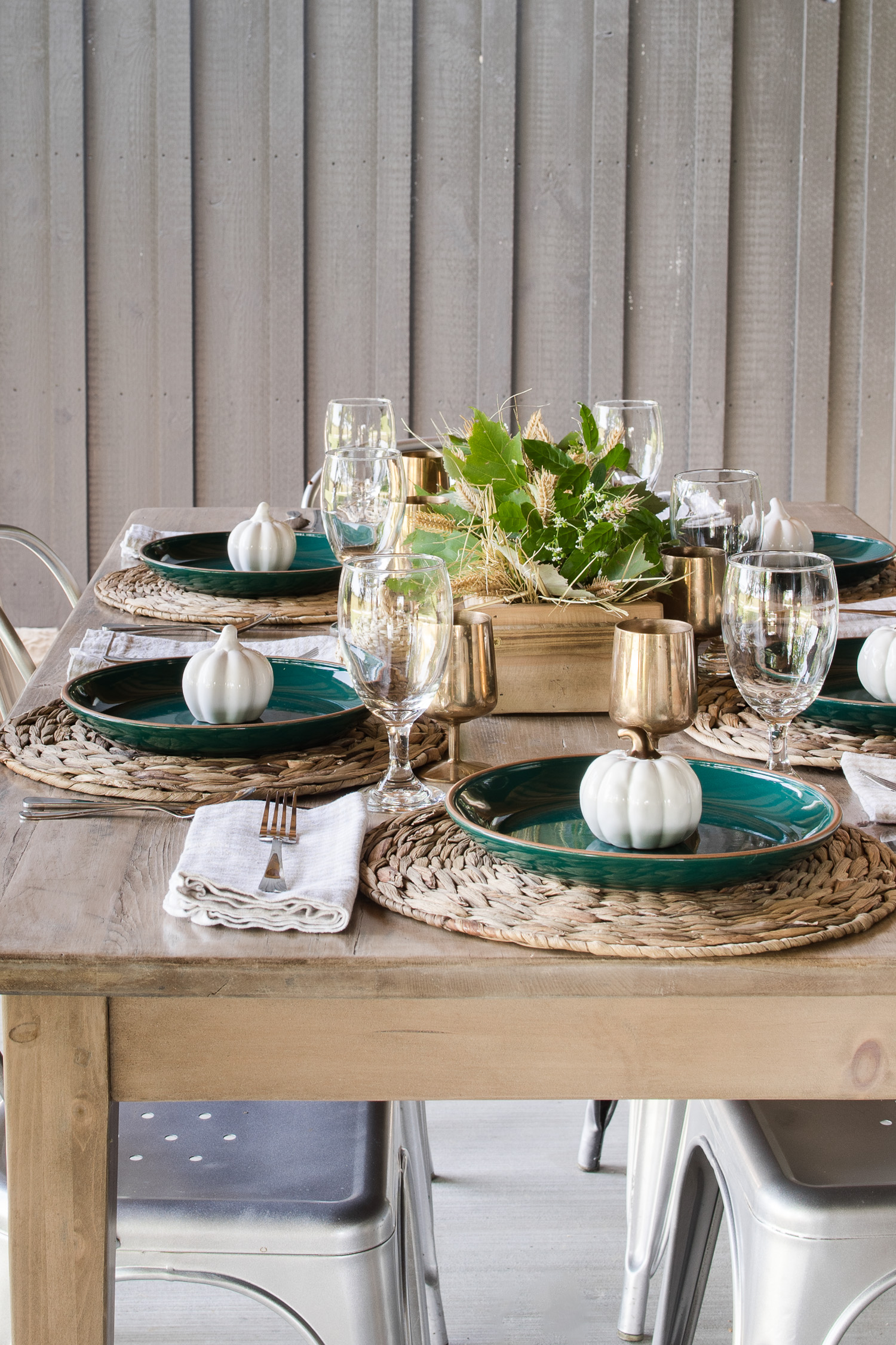 fall tablescape on the outdoor back patio with green plates, pumpkins and brass goblets. With a wooden centerpiece with green leaves and wheat stems