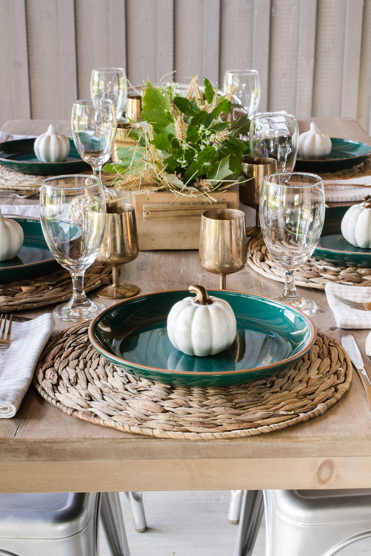 fall tablescape on the outdoor back patio with green plates, pumpkins and brass goblets. With a wooden centerpiece with green leaves and wheat stems